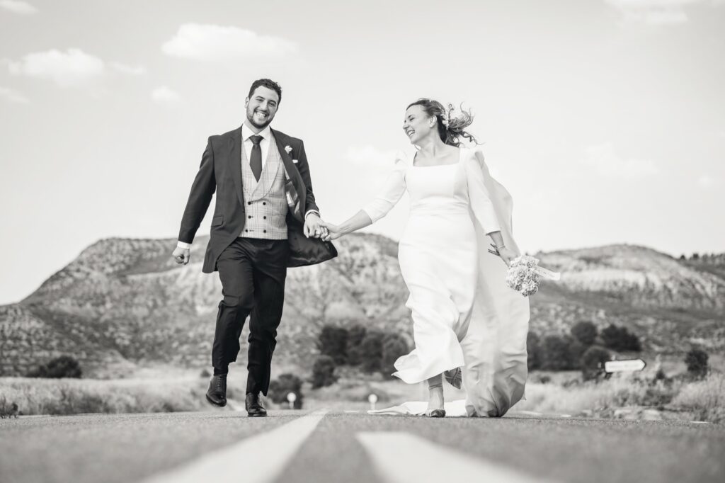 Fotos de boda divertidas: los novios corren de la mano por una carretera, en blanco y negro